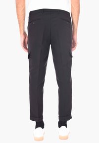 Uomo che indossa pantaloni cargo aderenti neri con tasche posteriori con bottone e polsini, abbinati a sneakers bianche e calze nere, in piedi rivolto verso l'indietro.