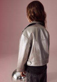Silverfärgad metallisk fake läderjacka med krage, texturerad yta och kort design. Modellen står bredvid en discokula på en rosa bakgrund.