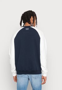 Marineblaues und weißes Sweatshirt mit Rundhalsausschnitt und Raglan-Design, aus Baumwollmaterial, mit gerippten Bündchen und einem kleinen Logo-Tag am hinteren Halsauschnitt.