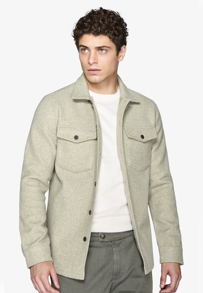 Lys grøn uldjakke med krave, frontknaplukning, to brystlommer og tekstureret stof, kombineret med en hvid sweater og grønne bukser.