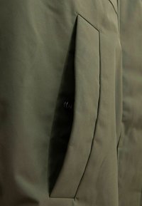 Tissu de veste vert olive présentant une grande poche insérée avec une fermeture à bouton, texture lisse et détails de couture robustes.