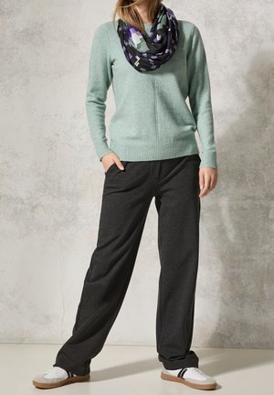 Personne portant un pull vert clair, une écharpe infinie noire à motifs, un pantalon noir ample et des baskets blanches, debout contre un mur texturé.