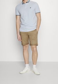 Světlomodré polo tričko s tmavým límcem, hnědé krátké kalhoty, bílé tenisky a viditelné logo na tričku. Jednoduchý neformální komplet.