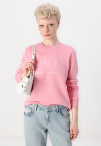 Light pink sweatshirt with embroidered logo, round neck, and ribbed cuffs, paired with light blue jeans. Model holds a small, white bag.