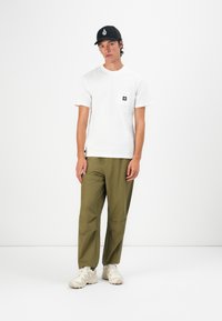 Young man wearing a white T-shirt with chest pocket, olive green pants, beige sneakers, and a black cap standing against white background.