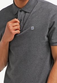 Ciemnoszara koszulka polo z klasycznym kołnierzykiem, trzema guzikami i małym niebieskim logo po lewej stronie na piersi. Wykonana z miękkiego, oddychającego materiału.