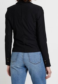 Černý sako s pruhovaným vzorem a strukturovaným střihem, se zapínáním na klopu a knoflíky na manžetách, spárované se světle modrými džíny.