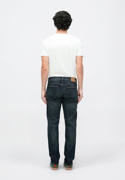 Hombre con cabello rizado negro que lleva una camiseta blanca de manga corta sencilla, jeans azul oscuro y zapatos multicolores, de pie mirando hacia atrás.