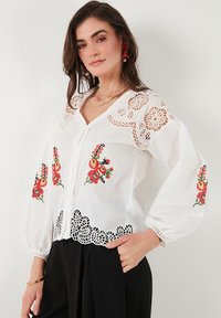 White blouse with intricate lace detail on shoulders and hem, featuring colorful floral embroidery on the front and sleeves.