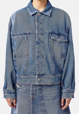 Person iført en blå denimjakke med knapgylpede brystlommer og matchende denimskørt, stående mod en ensfarvet baggrund.
