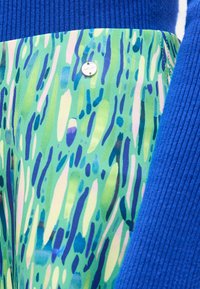 Φωτεινό μπλε πουλόβερ με ριγέ υφή δίπλα σε υφασμάτινο σχέδιο σε αποχρώσεις του πράσινου, μπλε και λευκού με μια μικρή στρογγυλή μεταλλική ετικέτα προσκολλημένη.