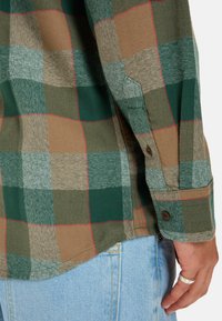 Camisa de cuadros en tonos verdes, marrones y grises con una textura de espiga. Presenta puños abotonados y un dobladillo curvado. Jeans de mezclilla claros visibles.