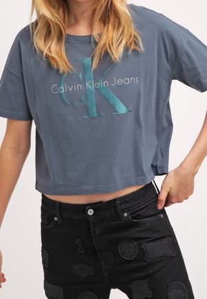 Osoba nosząca luźny szary crop top Calvin Klein Jeans i czarne jeansy z naszywkami, trzyma jedną ręką pas spodni.