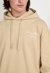 Beige huvtröja med avslappnad passform. Har en fram-logo i vit text, dragsko och mjuk bomullstextur.