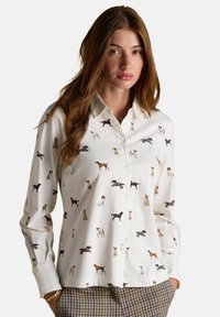 Žena s dlhými hnedými vlasmi oblečená v bielom košeli na gombíky s viacfarebnými ilustráciami psov a károvanými nohavicami, ręce v vreckách.