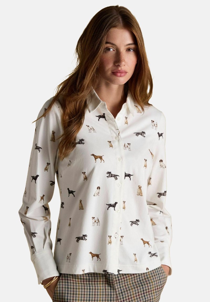 Žena s dlhými hnedými vlasmi oblečená v bielom košeli na gombíky s viacfarebnými ilustráciami psov a károvanými nohavicami, ręce v vreckách.