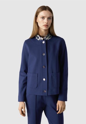 Frau trägt eine marineblaue, geknöpfte Jacke mit zwei Vorderentaschen über einem Hemd mit gemustertem Kragen, kombiniert mit passenden marineblauen Hosen.