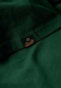 Tessuto verde scuro con una texture liscia, con un'etichetta nera con scritta dorata che recita "Superdry" alla cucitura.