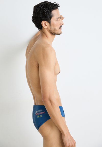 Blaue Schwimmbriefe aus glattem Stoff, mit mehrfarbigem gesticktem Text auf der Rückseite. Entworfen für eine enge Passform und minimalen Bedeckungsstil.