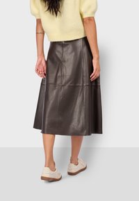 Falda midi de cuero marrón con cintura ajustada y forma acampanada. Presenta una textura suave y detalles de paneles geométricos. Llevada con zapatillas ligeras.