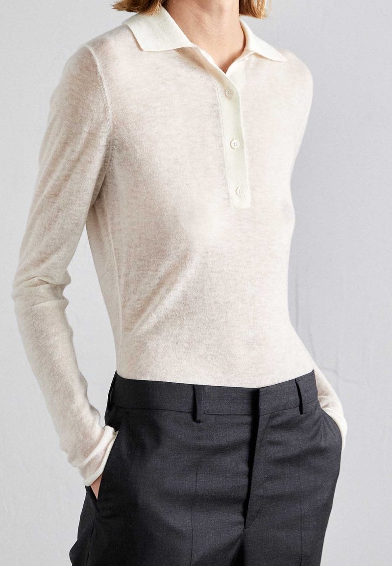 Polo a maniche lunghe di un beige chiaro, con pattina a bottoni, texture morbida e vestibilità casual, abbinato a pantaloni dark sartoriali.