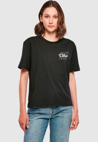 T-shirt en coton noir avec des manches courtes, un col rond et un motif sur le devant avec le texte "SEULES DES BONNES ONDES" en blanc.