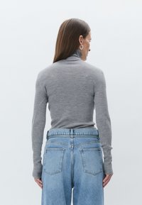 Kvinne med rett brunt hår iført grå ribbestrikket høyhalset genser og lys blå denim jeans med høy midje, stående med ryggen mot betrakteren.