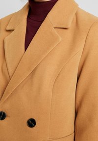 Kabát v barvě velbloudí s dvojitým zapínáním a velkými černými knoflíky, ostrými klopami a texturovanou tkaninou. Obsahuje přiléhavý siluetu a dobře zpracované švy.
