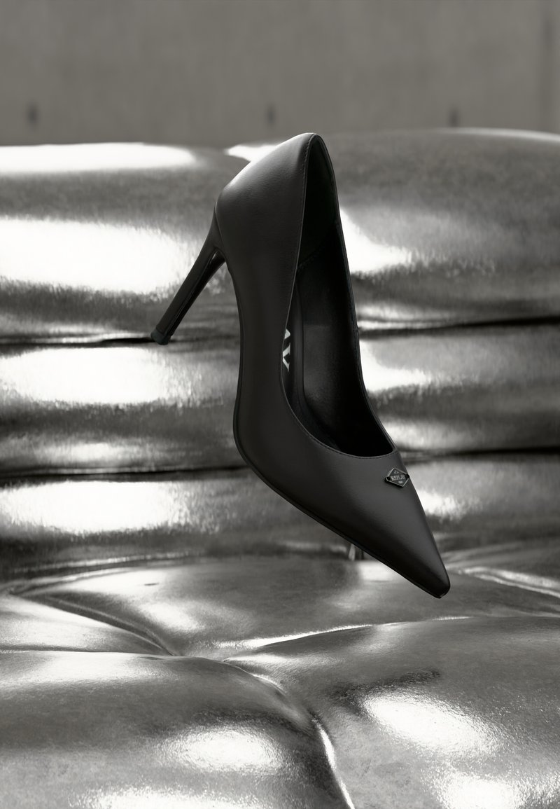 Μαύρο δερμάτινο παπούτσι με ψηλό τακούνι και μυτερή μύτη, με μεταλλική λεπτομέρεια στην περιοχή του πεταλού. Το παπούτσι τοποθετείται πάνω σε γυαλιστερή ασημένια επιφάνεια.