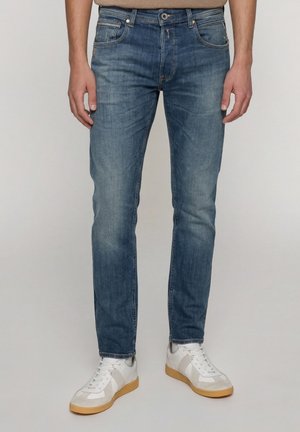 Mand iført slim fit blå jeans og hvide sneakers med gummisåler, stående mod en ensfarvet baggrund.