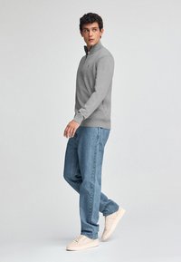 Grauer Reißverschluss-Pullover aus einem glatten Stoff, kombiniert mit blauen Jeans und hellen Sneakers. Das Modell steht im Profil.