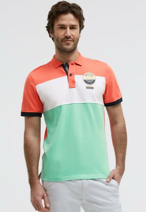 Mann trägt Polohemd mit kurzen Ärmeln in Orange, Weiß und Mintgrün, marineblaue Einfassung und segelthemenbezogenes Logo auf der Brust.