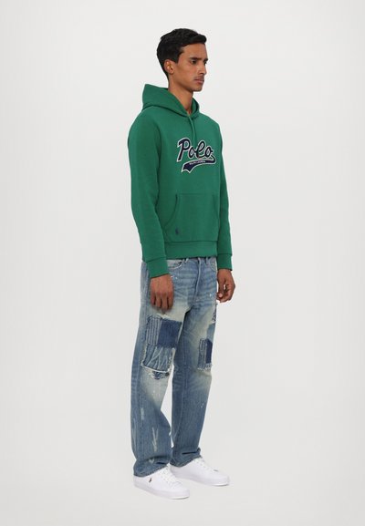 Felpa verde con cappuccio e logo "Polo", tasca a marsupio frontale, abbinata a jeans chiari strappati con toppe. Sneakers bianche.