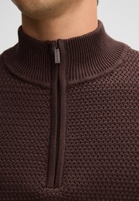 Brauner gestrickter Pullover mit hohem Kragen, ausgestattet mit einem halben Reißverschluss an der Vorderseite und einem strukturierten Muster, das sich durch den gesamten Stoff zieht.