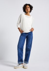 Witte gebreide trui met driekwartmouwen, gecombineerd met blauwe rechte jeans en beige sneakers met textuuraccenten.
