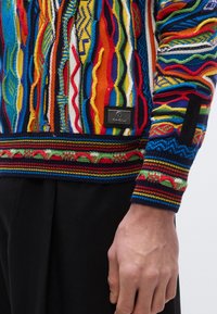 Pull tricot multicolore avec des motifs complexes, des rayures horizontales et un design texturé ; comprend une étiquette de marque noire sur le côté.