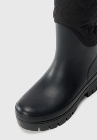 Bottes noires en caoutchouc jusqu'à la cheville avec une semelle large et texturée et un upper en tissu matelassé. Présente une finition lisse et un design à bout arrondi.