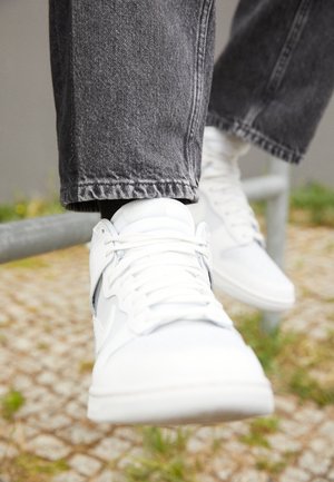 Nahaufnahme weißer Sneakers, getragen zu schwarzen Jeans, Füße ruhen auf einem Metallgeländer im Freien auf gepflasterter Fläche mit Grasflecken.