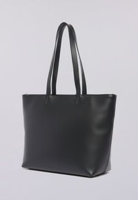 Černá kožená taška přes rameno s dlouhými dvojitými popruhy a zipovým uzávěrem, na jednolitém světlém šedém pozadí.