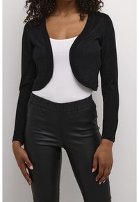 Černý krátký kardigan s mírně lesklým povrchem, s dlouhými rukávy a otevřeným předním dílem, v kombinaci s černými kalhotami z umělé kůže s vysokým pasem.