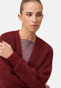 Bordowy, miękki kardigan z dekoltem w kształcie litery V. Głęboko dziany wzór i prążkowane mankiety. Pod spodem dopasowana, paskowana koszula w bordowym i białym kolorze.