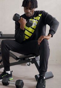 Felpa sportiva nera con strisce gialle, pantaloni della tuta neri e sneakers nere con accenti verdi. Tiene un manubrio nero.