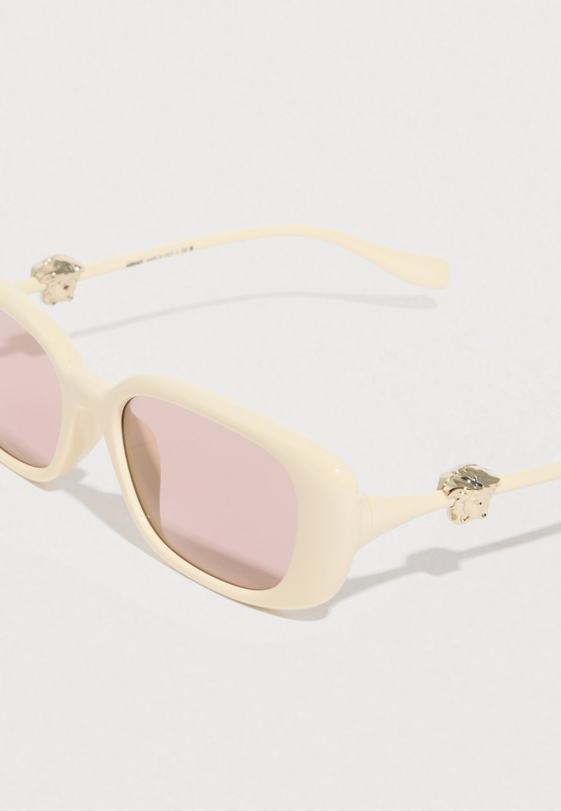 Rektangulære solbriller i cremefarve med pinke linser og gyldne løvehoveddekorationer på siderne mod en hvid baggrund.