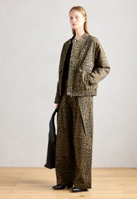 Leoparda raksta jaka un platību bikses brūnā un melnā audumā, kombinētas ar melnu somu un smailām kurpēm. Zem tā ir vienkrāsaina krekls.