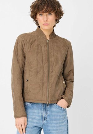 Junge Person mit kurzen lockigen Haaren, trägt eine braune gesteppte Jacke mit Reißverschluss und hellblaue Jeans, Hand in der Tasche, neutraler Hintergrund.