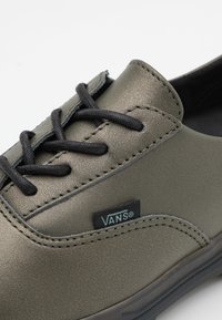 Shoe sintético verde-oliva com cadarços e costura pretos, apresentando um pequeno rótulo preto da Vans na lateral, bico arredondado e acabamento suave.