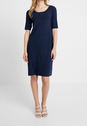 Femme portant une robe bleu marine longueur genou à manches courtes et des sandales blanches à talons hauts à lanières, debout devant un fond blanc uni.