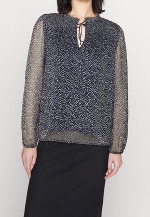 Blouse marine avec motif floral blanc, manches longues semi-transparentes et détail noué au col. Tissu doux avec une coupe décontractée, associée à une jupe noire.