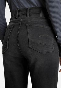 Vista trasera de una persona que lleva jeans de mezclilla negros de talle alto y una camiseta de manga larga de canalé oscura, con las manos relajadas a los lados.