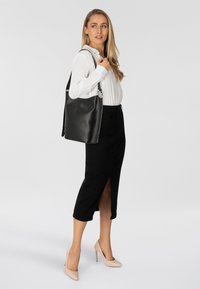 Borsa a mano nera in pelle dal tessuto liscio, con finiture in argento e una tracolla singola, indossata da una modella con una camicia bianca.